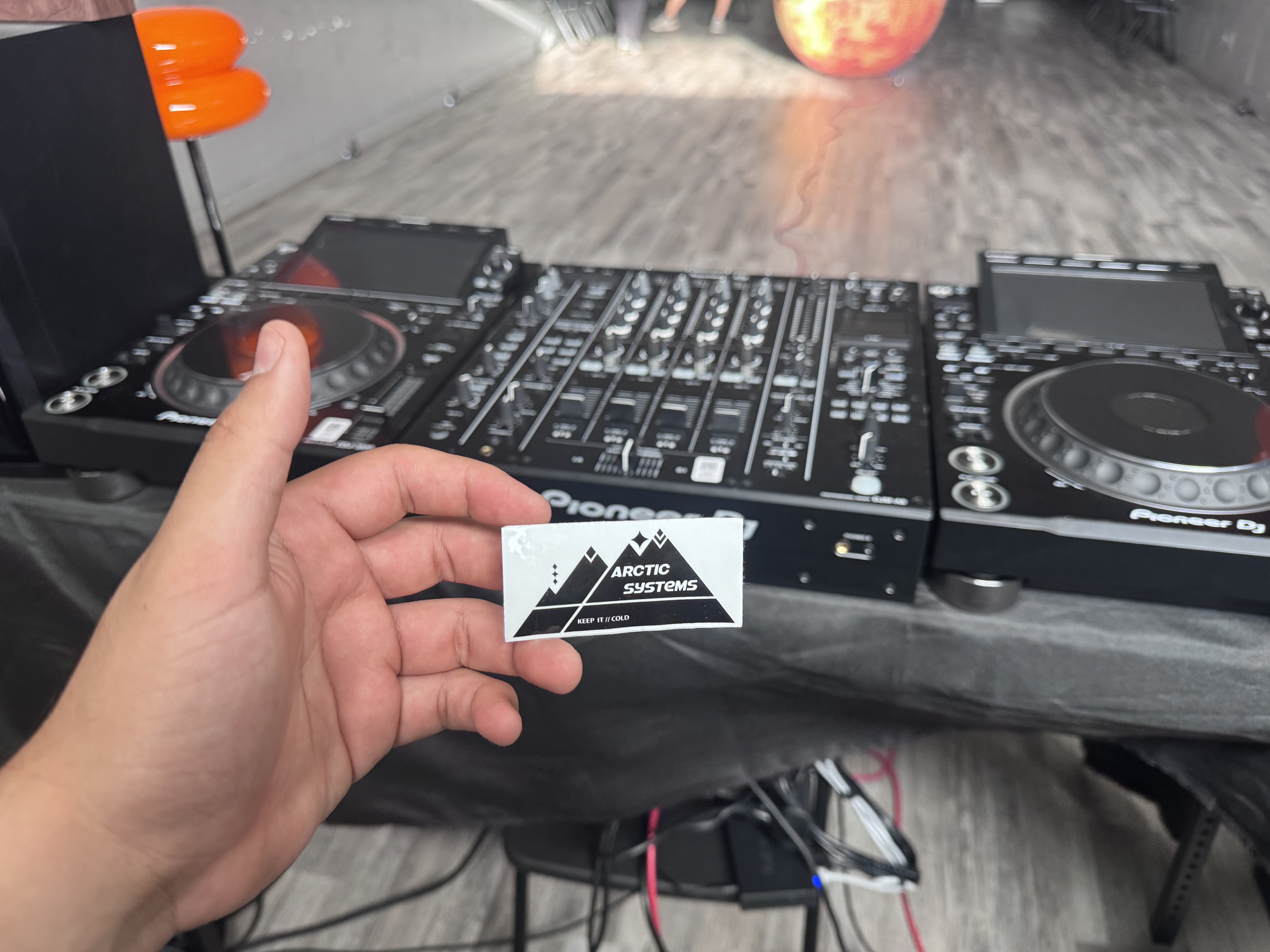 A hand holding an Arctic Systems mountain-logo sticker in front of Pioneer DJ decks